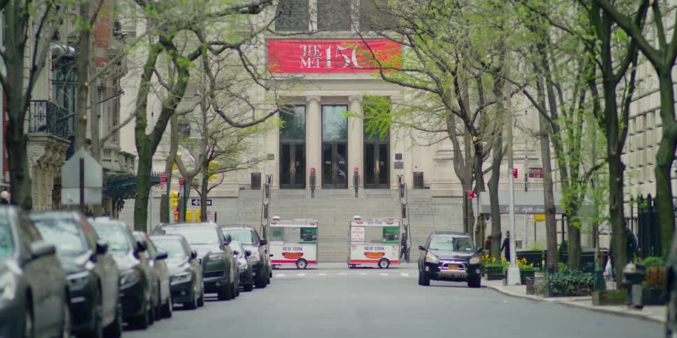 Two empty and unmanned hot dog carts parked near the steps to the metropolitan museum as a few pedestrians walk by