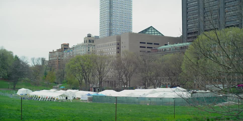 Several white tents are set up on a lawn behind several tall buildings in a city