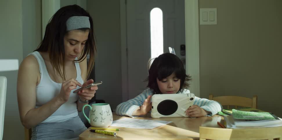 A woman sitting at a table using a smart phone while a girl uses a tablet