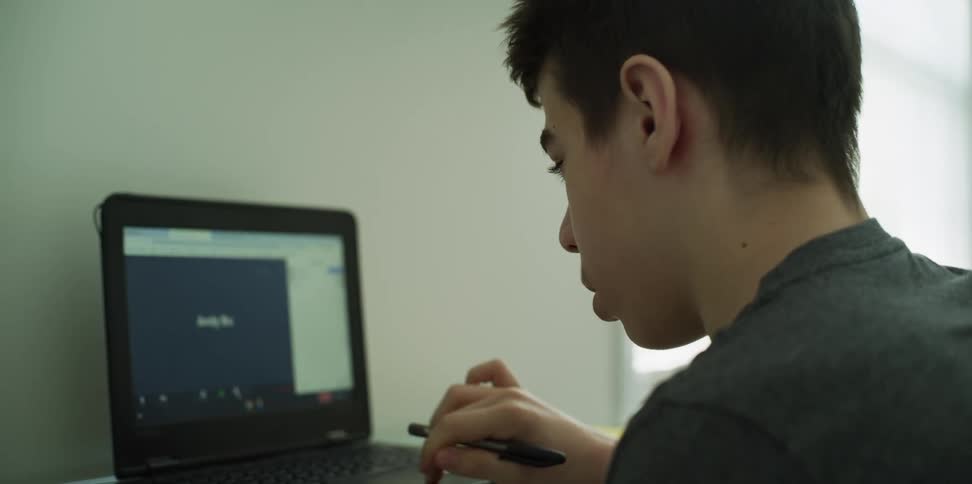 A boy with a pen in his hand typing on a laptop