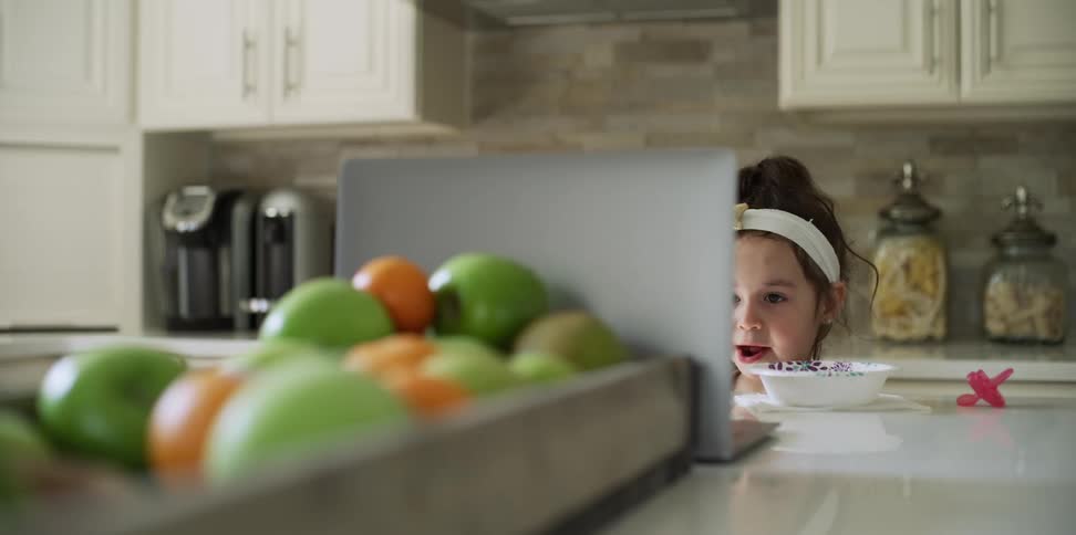 Female child stands in front of the kitchen island watching an open laptop while chewing on food from a bowl on the counter