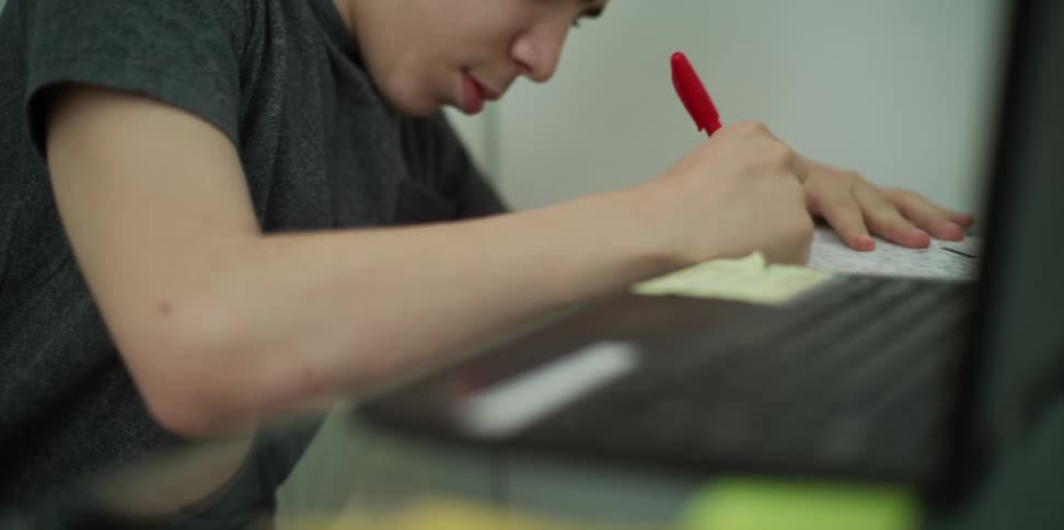 A teenage boy uncaps a pen to write something down as he sits at his desk with a laptop
