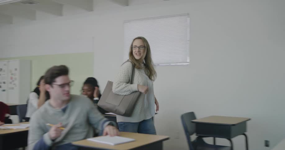 A teacher enters a classroom from the back and waves and greets the students as she walks up to the front