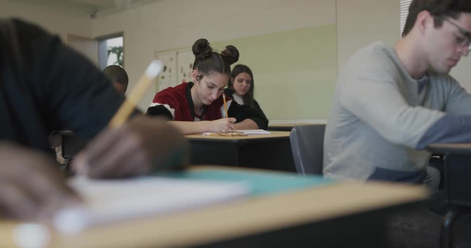 Students sit at their desks in a classroom writing in notebooks with pencils