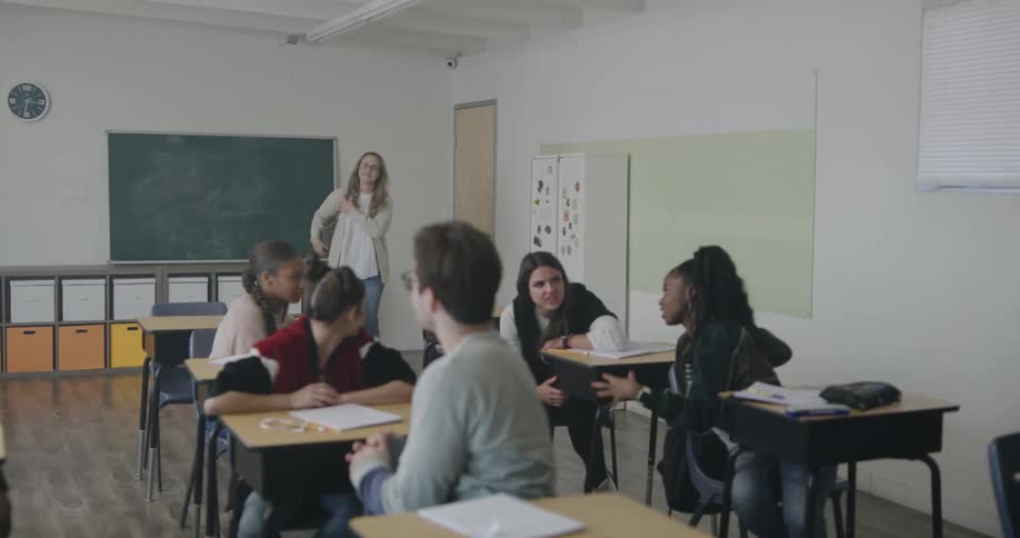 Students sitting in a classroom while the teacher enters and walks towards the front