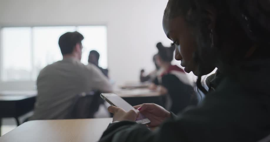 A teenage student sits and types on a smartphone while other students sit at their desks and talk to each other