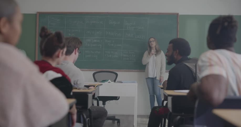A classroom where students are sitting while the teacher is standing by the blackboard teaching
