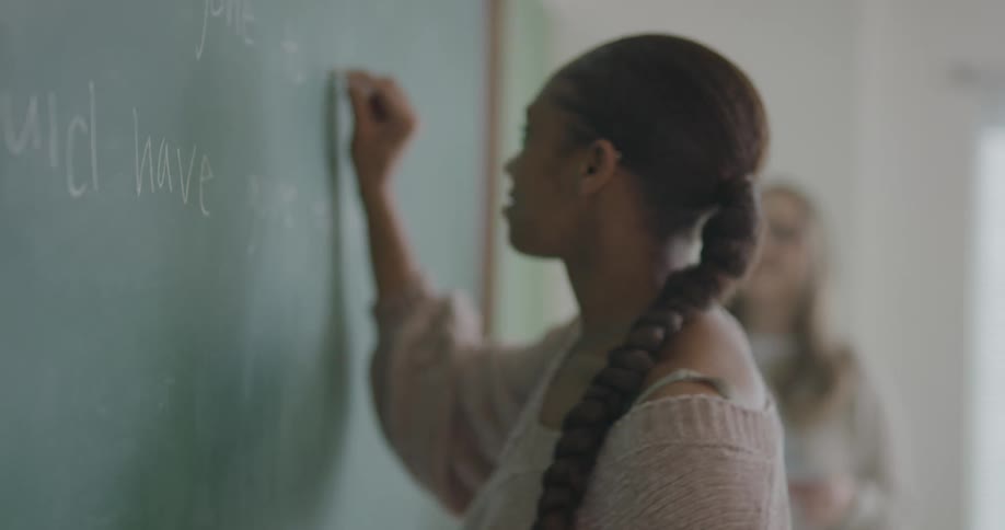 A girl writing on the blackboard while the teacher talks to the class