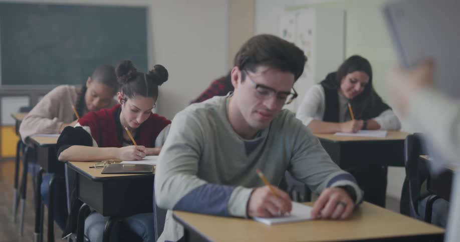 Students in a classroom sit at their desk listening to the teacher and writing in notebooks with pencils