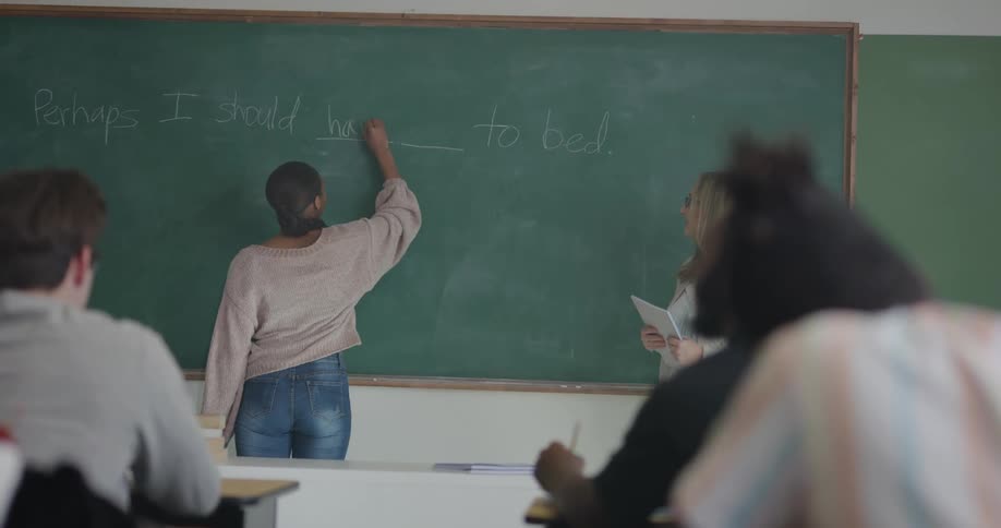 A teacher standing at the board has a student fill in the missing words to complete a sentence in front of the class