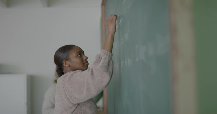 A student writes on a chalkboard as the teacher instructs her
