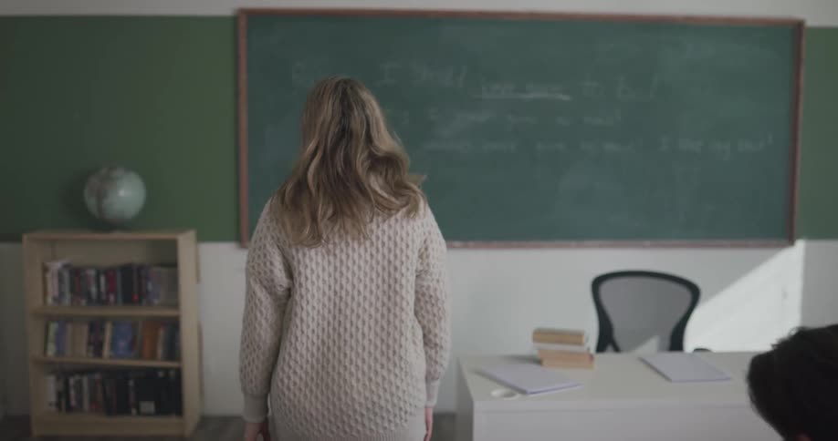 A teacher walks to the front of the classroom and sits at her desk