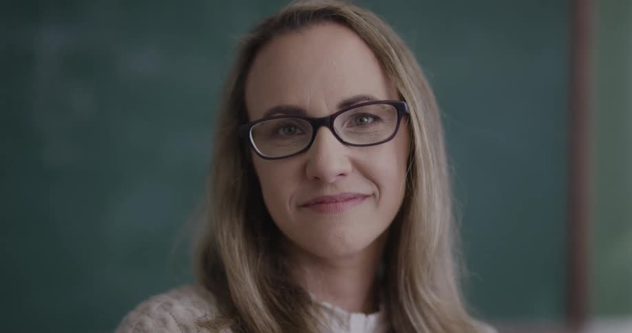 A teacher stands near the green chalkboards and looks and smiles at the class