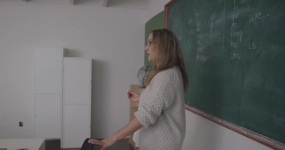 Blonde female teacher writes on the blackboard and teaches the class