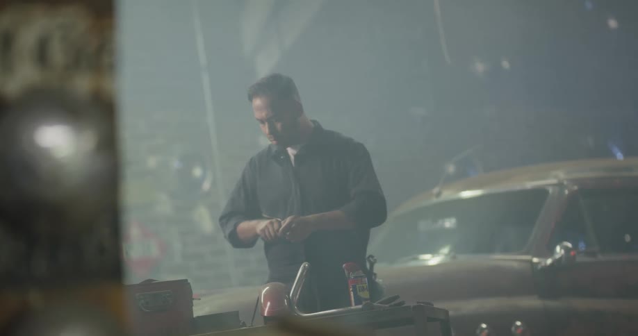 A mechanic in a garage adjust a part at his workbench then turns back to a vintage car