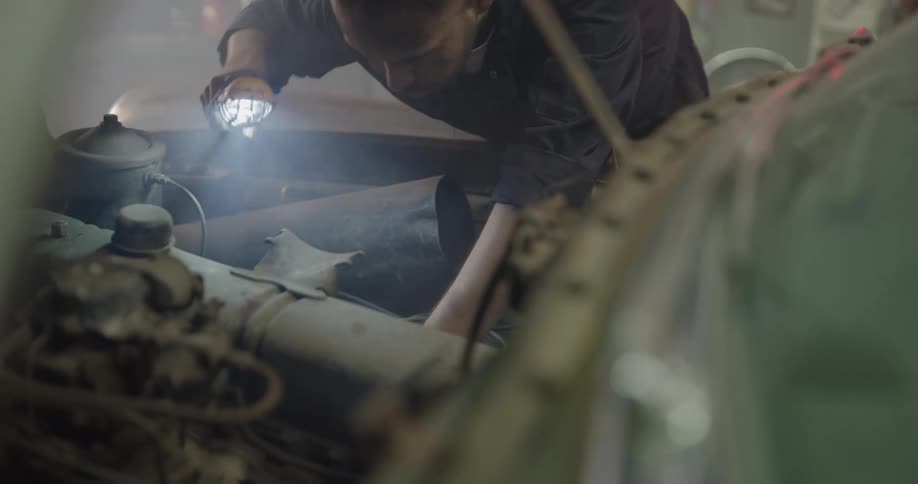 A mechanic holds a work light as he examines under the hood of an old rusty car