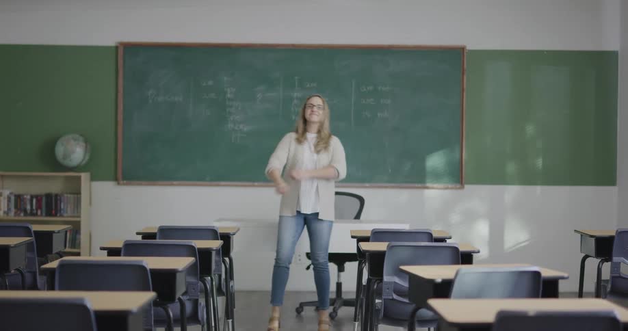 A teacher walking in front of her desk then standing and crossing her hands