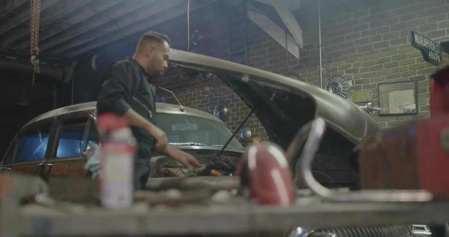 A mechanic in a garage works under the hood of a vintage car then walks over to his tool box
