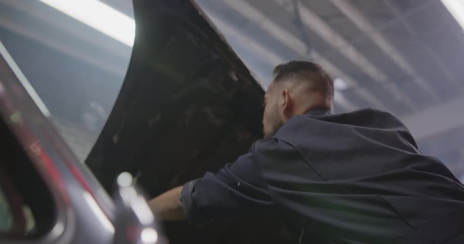 A mechanic pops the hood of a rusty car and bend over to take a look at the engine