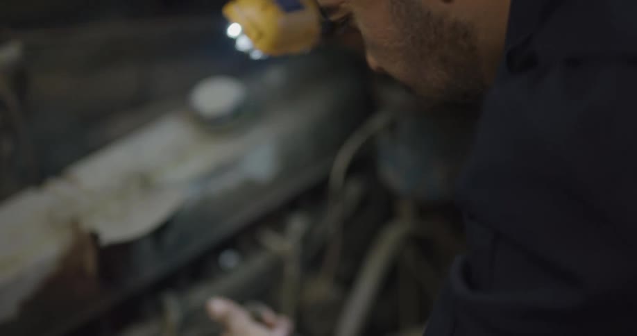 A mechanic shining a light and looking at wires underneath a car hood