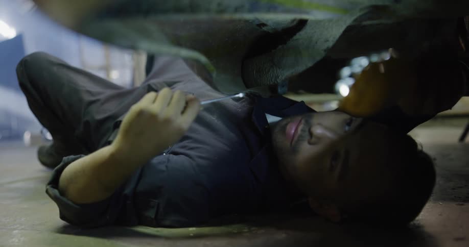 A mechanic on his back in a garage holding up a light and using a wrench to repair a car