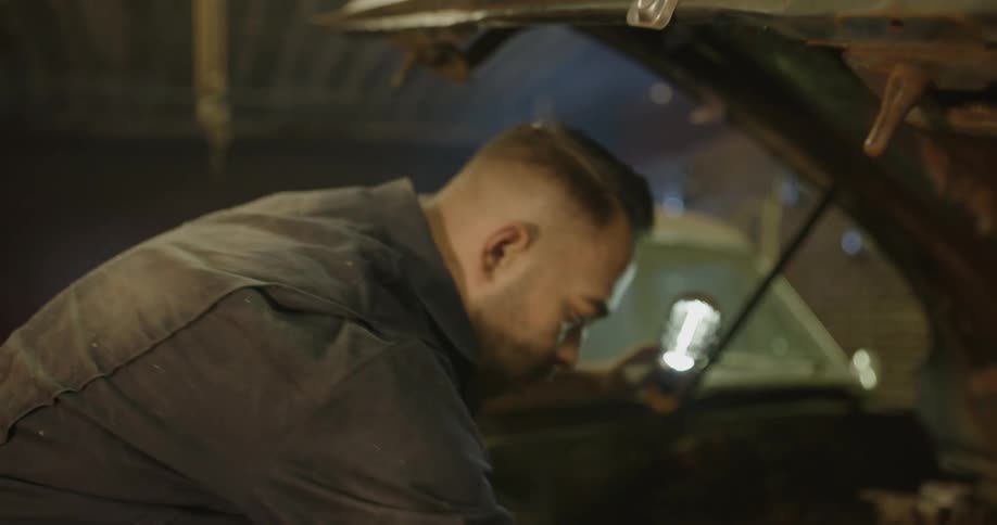 A mechanic holding a work light works on an old car engine in a garage