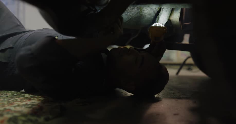 A mechanic on his back under a car holding a work light and a tool making a repair in a garage