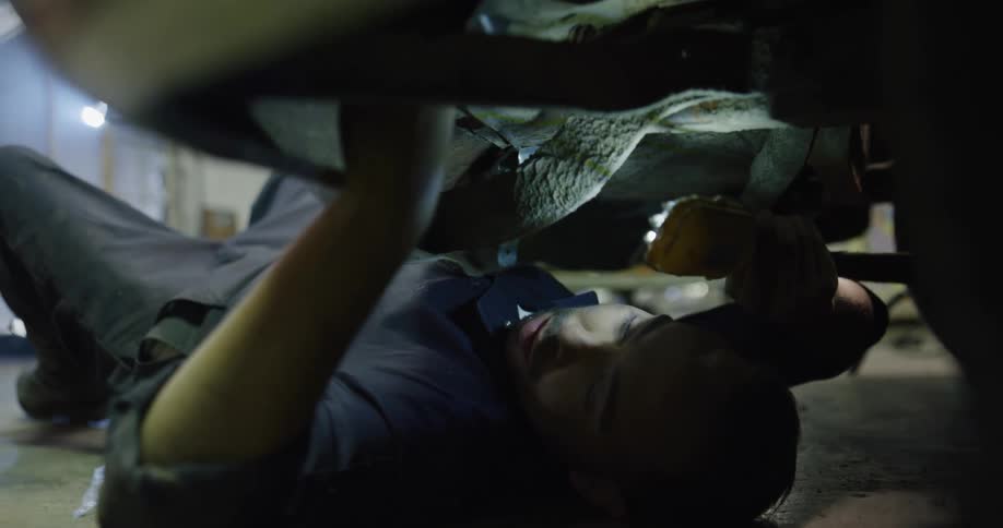 Car mechanic lies on back under car using light and wrench
