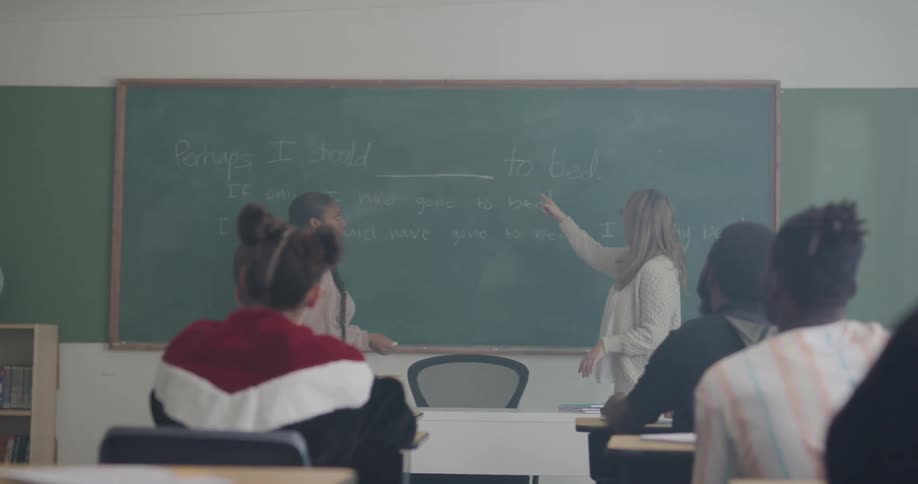 Students raise their hands in a classroom and the teacher chooses one student to write on the board