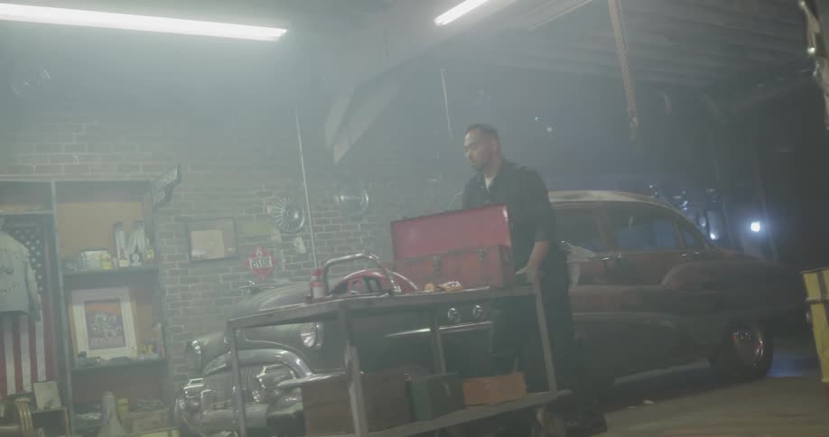Car mechanic pushes cart with toolbox past vintage car in dimly lit garage