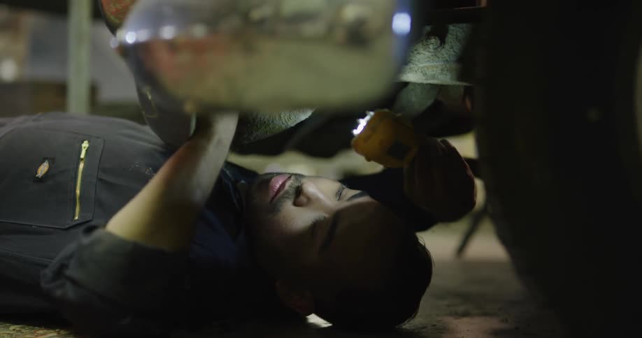A male mechanic lays on the floor with a flashlight looking under a car