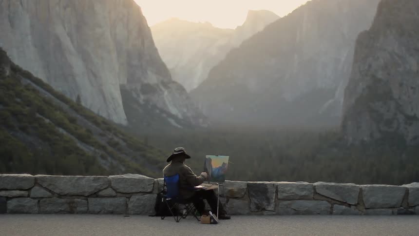 A man sitting next to a short, stone wall painting a picture of the mountain view before them