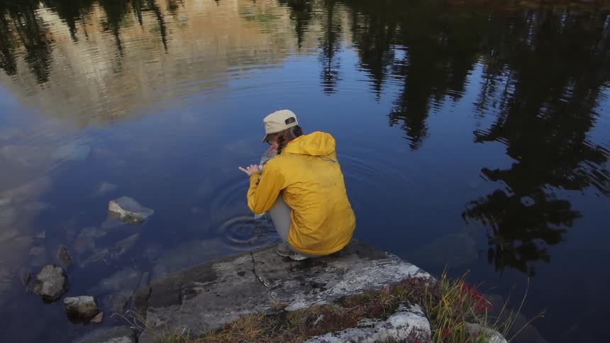 A woman kneels by a lake or pond and pours water into a container