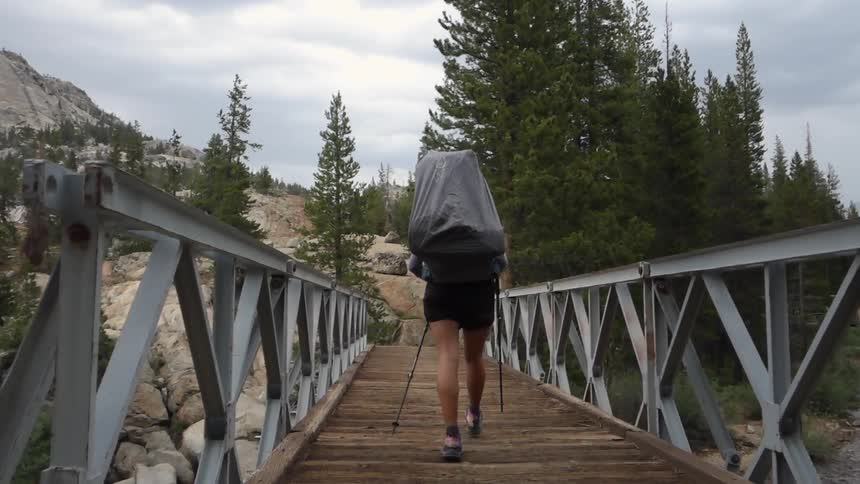 A hiker, carrying a large backpack, crossing a bridge suspended over a river in the mountains on a cloudy day