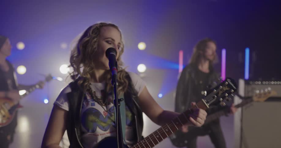 A female band leader sings in a microphone and plays the guitar