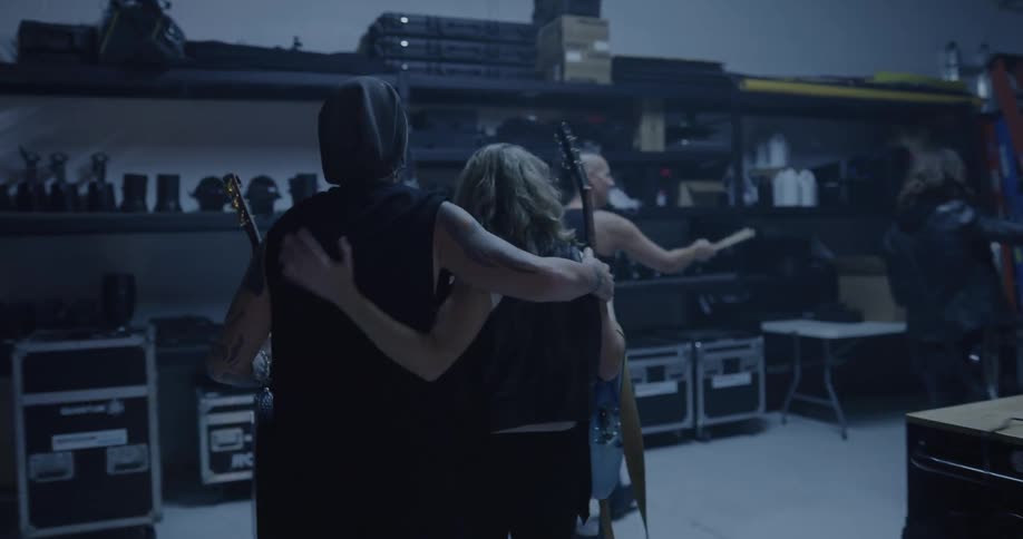 A man carrying a guitar hugging a woman carrying a guitar while walking down a backstage hallway
