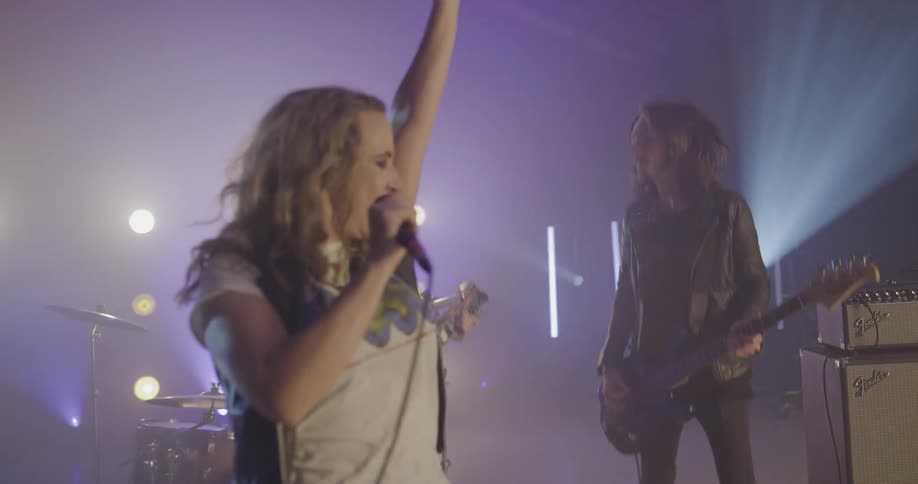 A female lead band member sings into a microphone while the other band mates play guitar