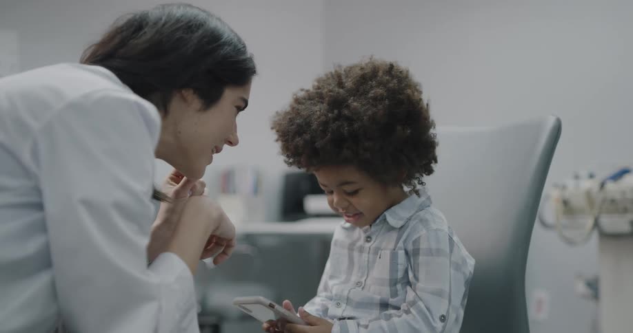 A doctor makes her young patient smile as he holds a smartphone