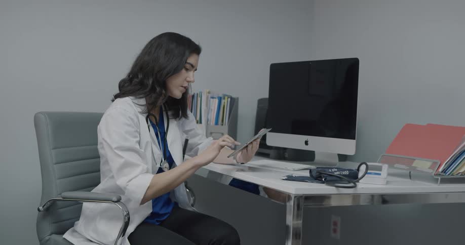 A doctor sits at a desk in an office wearing a lab coat and tapping and scrolling on a tablet