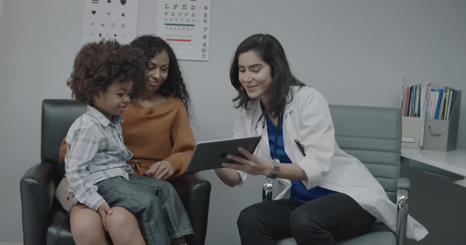 A little boy sits on his moms lap as a doctor talks to them and shows them things on an ipad