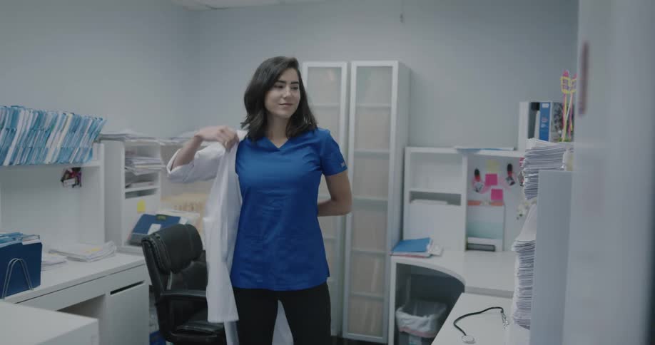 A doctor standing in an office picks up a white lab coat and puts it on with a smile on her face