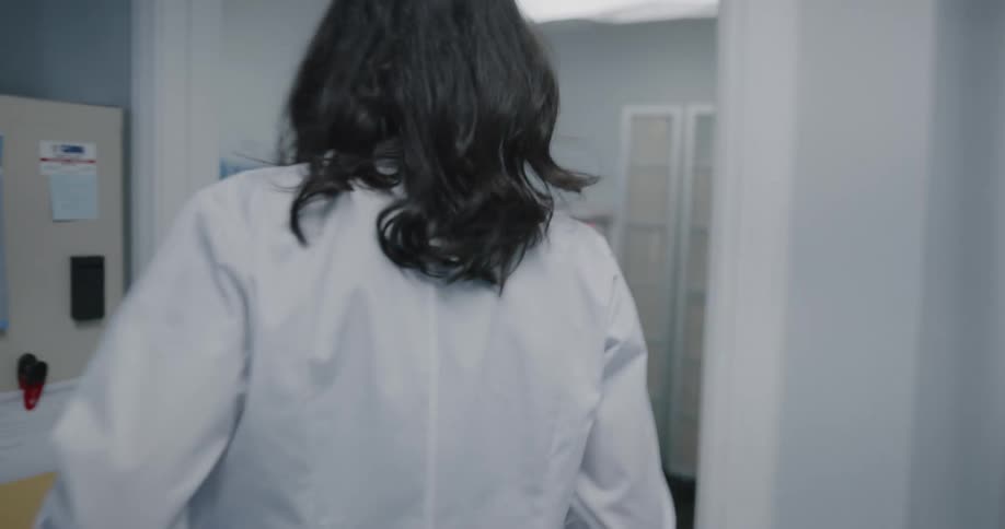 A doctor walks to her office and takes her lab coat off