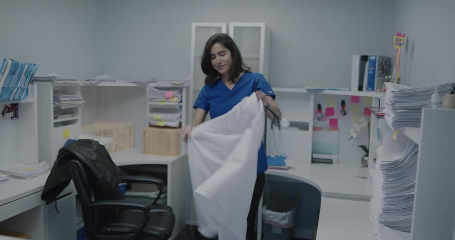 A doctor walks in to her office and takes her lab coat off and puts her stethoscope down