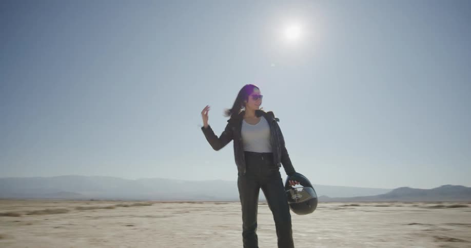 A woman standing in the desert holding a helmet with the sky in the background