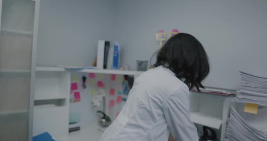 A female doctor puts on her lab coat in her office and walks out of the door