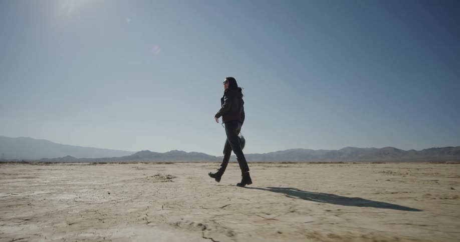 Woman wearing motorcycle leathers and carrying helmet walks across desert near foggy mountains