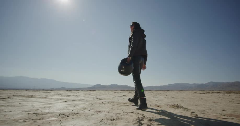 A woman holding a helmet walks across a desert