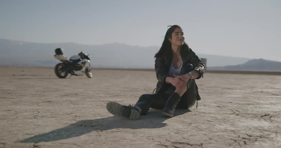 A woman sits on the ground in the desert with her motorcycle in the background