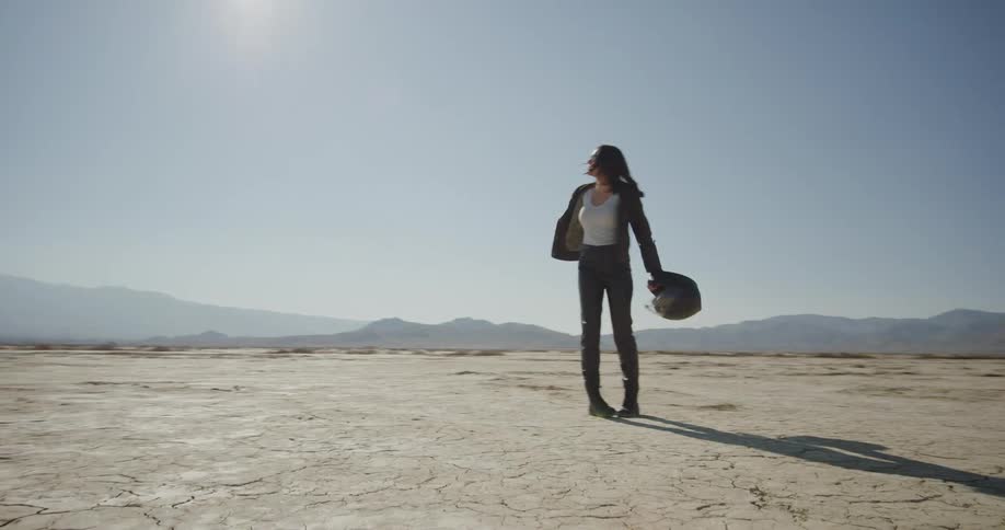 A woman holding a crash helmet looks and spins around in the desert