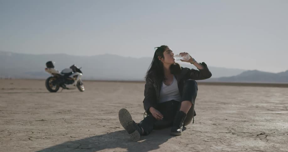 A woman sitting on the soil takes a drink from a water bottle